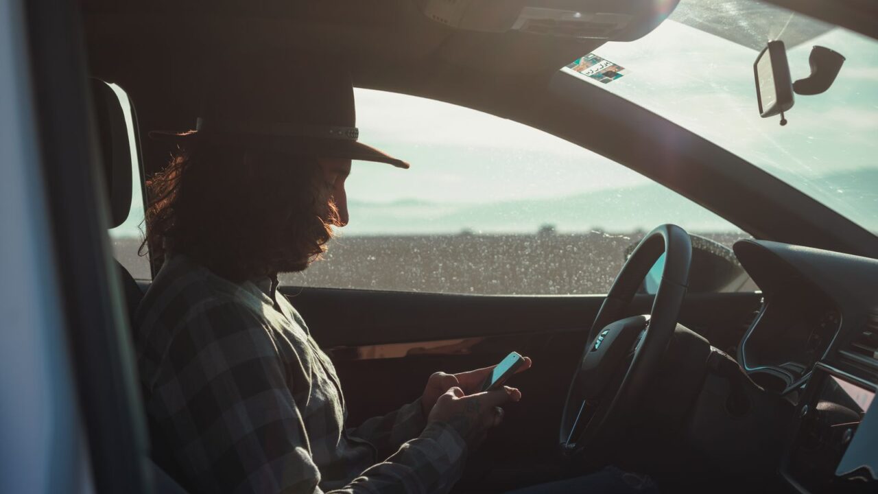 Homem dirigindo consulta o celular dentro do carro antes de sair, representando a checagem da validade da CNH