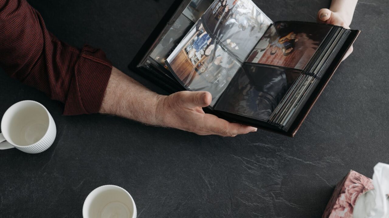 Mãos de duas pessoas segurando um álbum de fotos antigo sobre uma mesa, com xícaras de café ao lado