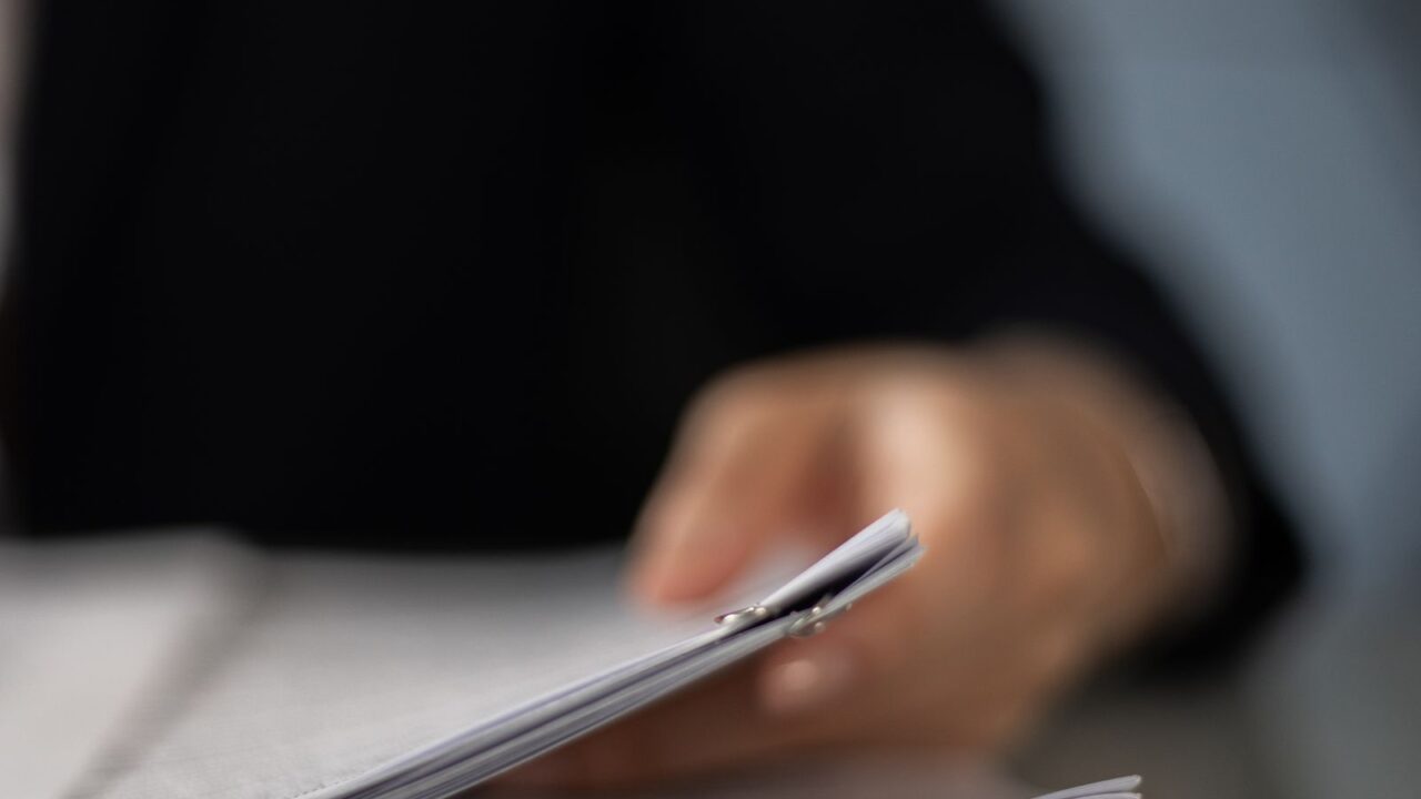 Mão desfocada segurando pilha de documentos sobre mesa de escritório, ilustrando regularização cadastral