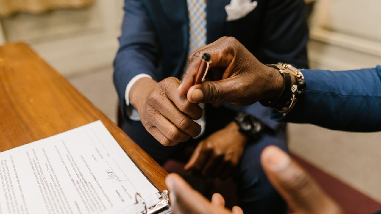 Dois homens de terno apertando as mãos sobre um documento assinado, representando o fechamento de um contrato empresarial
