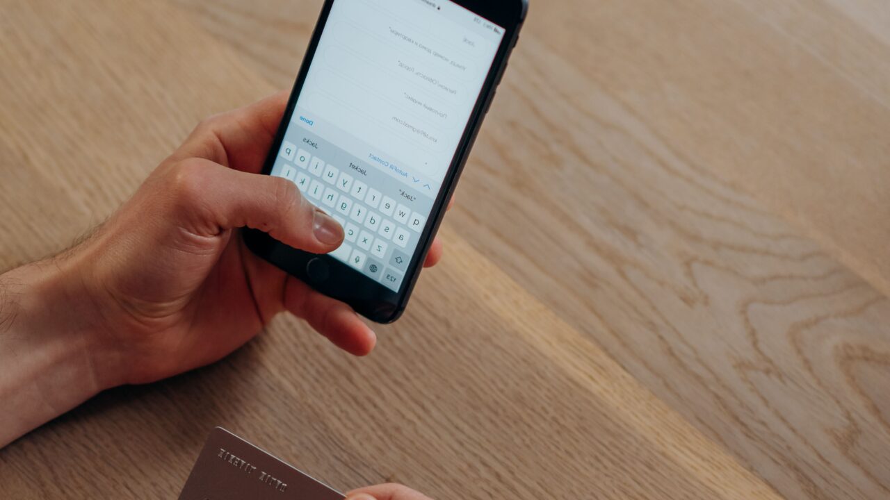Mãos segurando um celular com aplicativo de banco digital aberto e um cartão sobre uma mesa de madeira