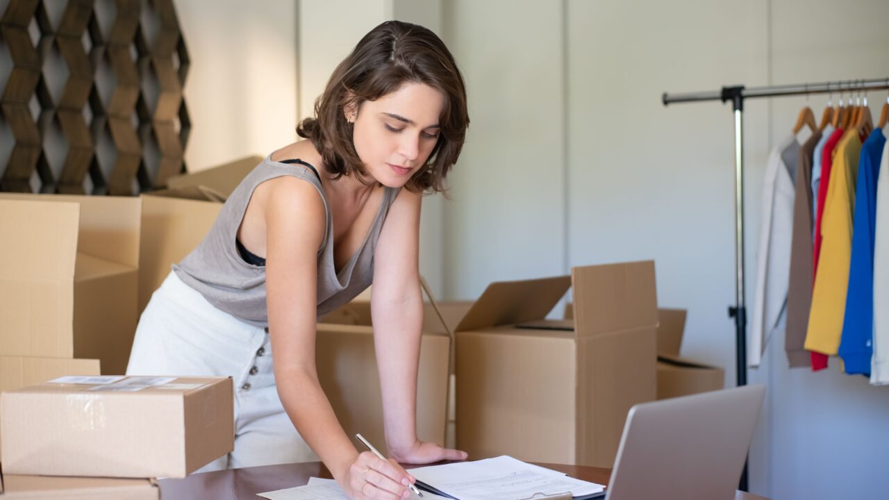 Mulher autônoma escrevendo anotações sobre uma mesa com caixas de papelão e um varal de roupas ao fundo